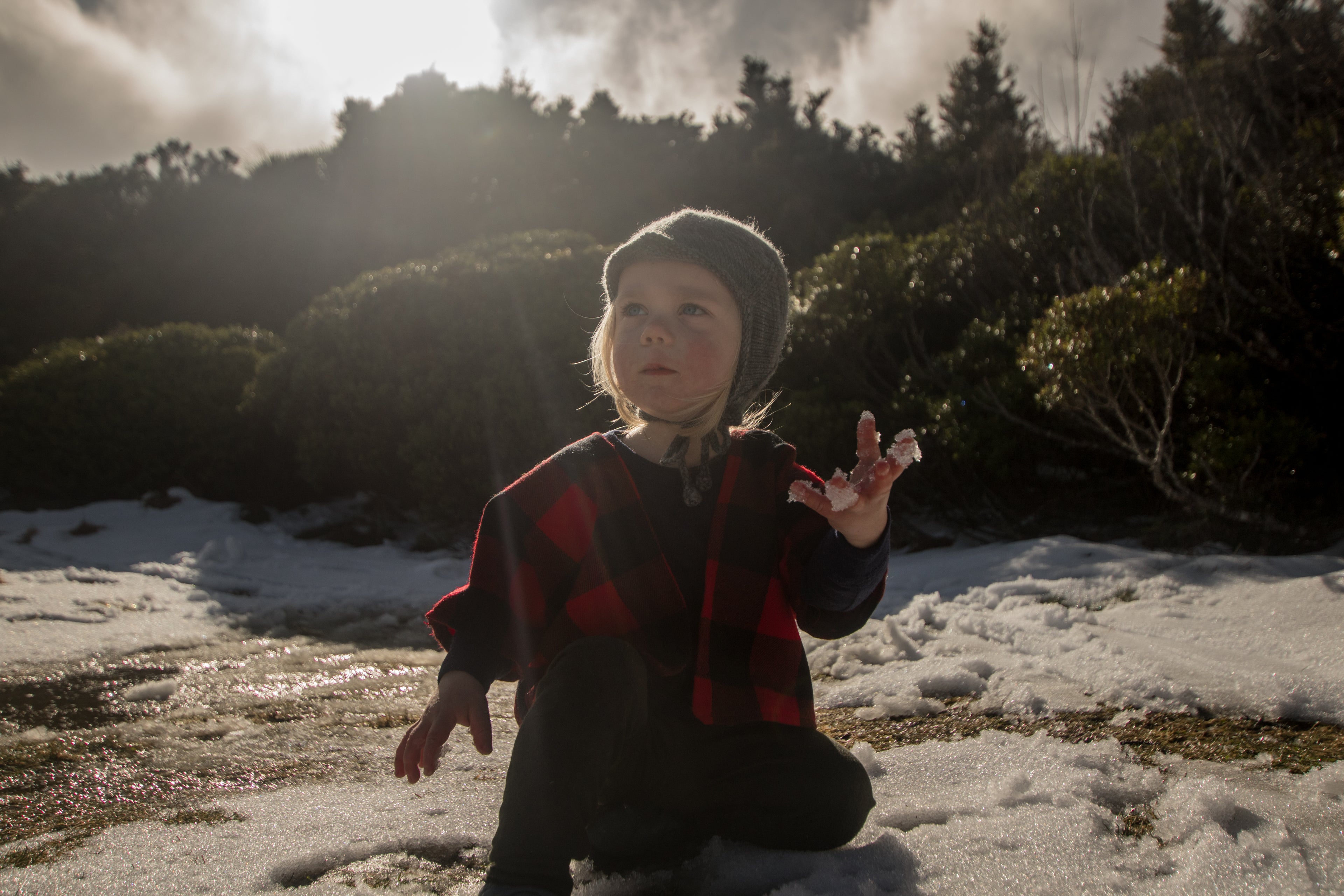Child in a red jacket and black pants standing in a snowy landscape with trees in the background.