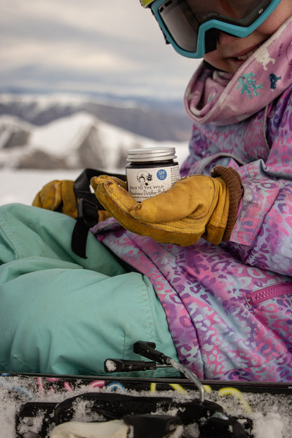 Person in winter gear holding a jar of cream with mountains in the background