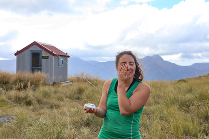 Woman in a green shirt standing in a grassy field with a small building and mountains in the background.